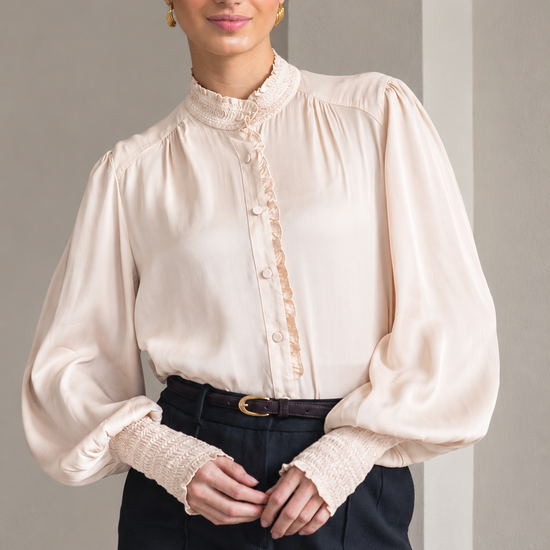 A woman modeling a cream-colored blouse with a high ruffled collar, gathered shoulders, and lace-trimmed cuffs, paired with dark trousers.