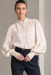 A woman modeling a cream-colored blouse with a high ruffled collar, gathered shoulders, and lace-trimmed cuffs, paired with dark trousers.