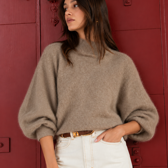 A woman wearing a fuzzy taupe-brown mock-neck bubble-sleeve sweater and white jeans, standing in front of a red wooden door.