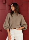 A woman wearing a fuzzy taupe-brown mock-neck bubble-sleeve sweater and white jeans, standing in front of a red wooden door.