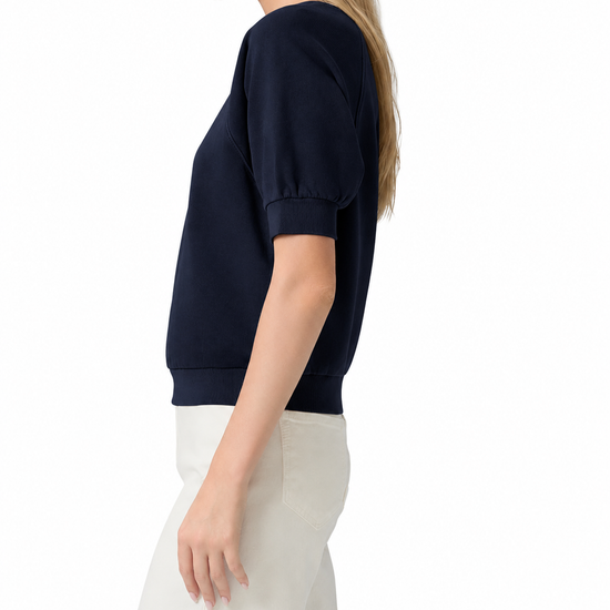 A woman shown in side profile wearing a navy blue short-sleeve top with puff sleeves and white pants, photographed against a clean white studio background.