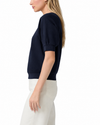 A woman shown in side profile wearing a navy blue short-sleeve top with puff sleeves and white pants, photographed against a clean white studio background.
