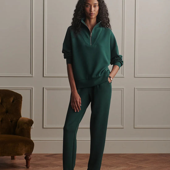 A woman stands in a room with paneled walls and wood flooring, wearing a matching dark green two-piece tracksuit with a half-zip pullover and straight-leg pants. A brown velvet armchair is visible on the left.