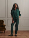 A woman stands in a room with paneled walls and wood flooring, wearing a matching dark green two-piece tracksuit with a half-zip pullover and straight-leg pants. A brown velvet armchair is visible on the left.