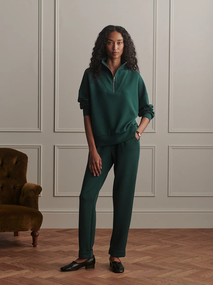 A woman stands in a room with paneled walls and wood flooring, wearing a matching dark green two-piece tracksuit with a half-zip pullover and straight-leg pants. A brown velvet armchair is visible on the left.