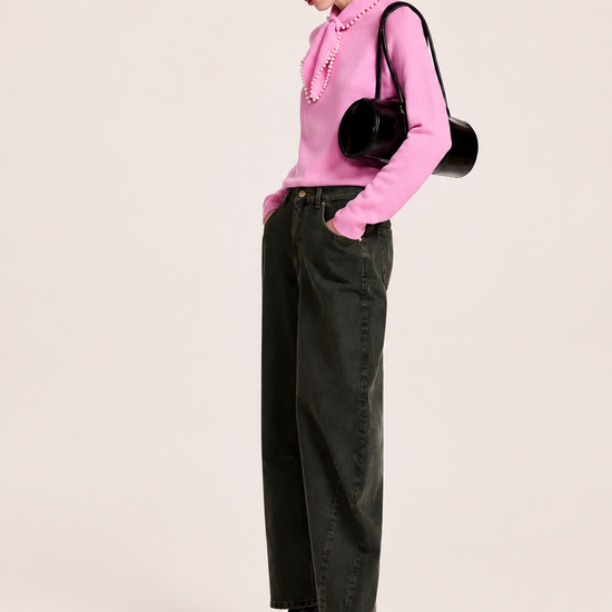 A model is shown from the side wearing a pink ruffled knit top, dark olive green wide-leg jeans, black patent leather boots, and a black baseball cap. She is carrying a black round shoulder bag against a solid light beige background.