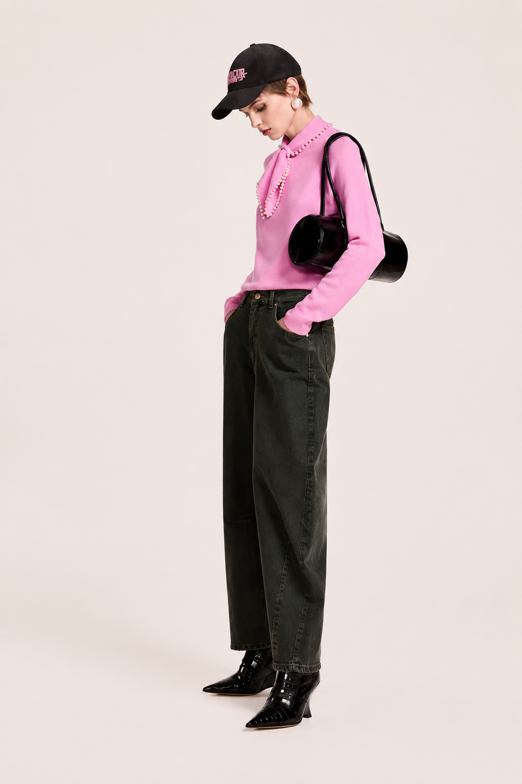 A model is shown from the side wearing a pink ruffled knit top, dark olive green wide-leg jeans, black patent leather boots, and a black baseball cap. She is carrying a black round shoulder bag against a solid light beige background.