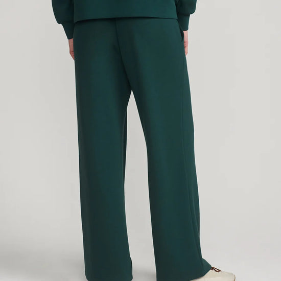 A person shown from the back wearing dark green wide-leg trousers and a matching dark green top, paired with off-white sneakers, against a light gray studio background.