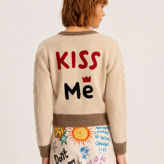 A model shown from the back wearing a cream-colored cable-knit sweater with 'KISS Me' written on the back in red and black, paired with a matching cream skirt featuring colorful doodle graphics.