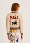 A model shown from the back wearing a cream-colored cable-knit sweater with 'KISS Me' written on the back in red and black, paired with a matching cream skirt featuring colorful doodle graphics.