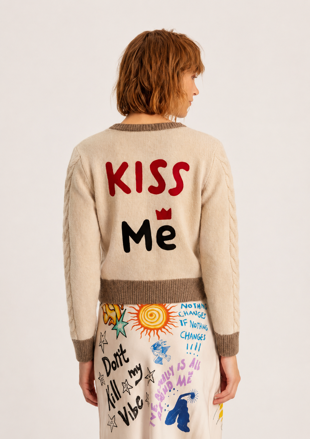 A model shown from the back wearing a cream-colored cable-knit sweater with 'KISS Me' written on the back in red and black, paired with a matching cream skirt featuring colorful doodle graphics.