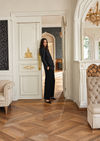 A woman wearing a black wide-leg blazer and matching wide-leg trousers stands in an elegant interior doorway with wood parquet flooring, cream walls, and gold decorative accents.