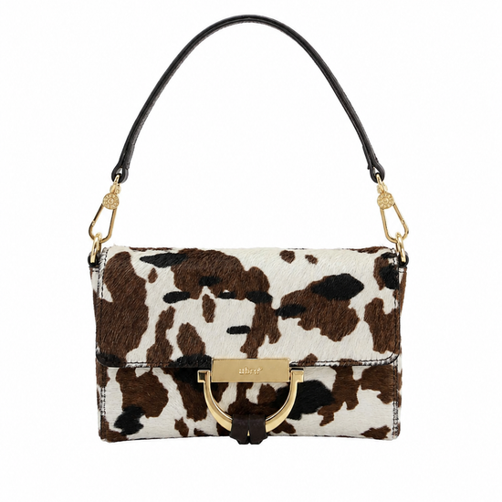A small wristlet clutch with a brown and white cow print pattern, a dark brown wrist strap, and gold-tone hardware, shown against a plain white background.
