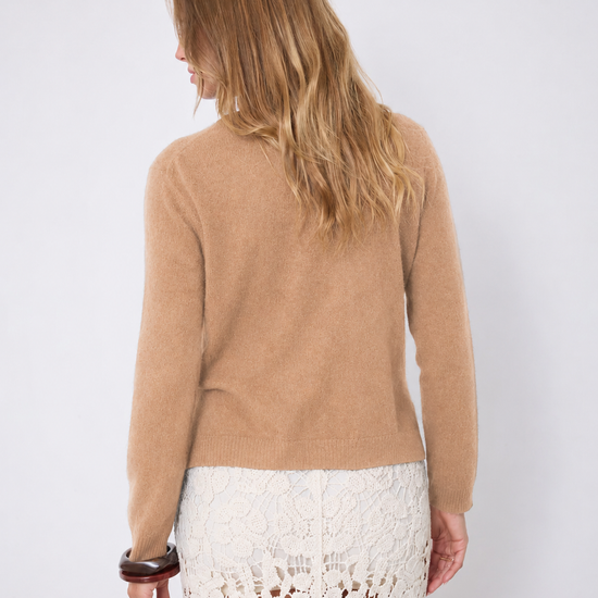 A woman shown wearing a cropped, long-sleeve knit cardigan in a warm camel tan color over a white lace skirt, set against a plain light gray studio background.