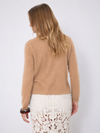 A woman shown wearing a cropped, long-sleeve knit cardigan in a warm camel tan color over a white lace skirt, set against a plain light gray studio background.