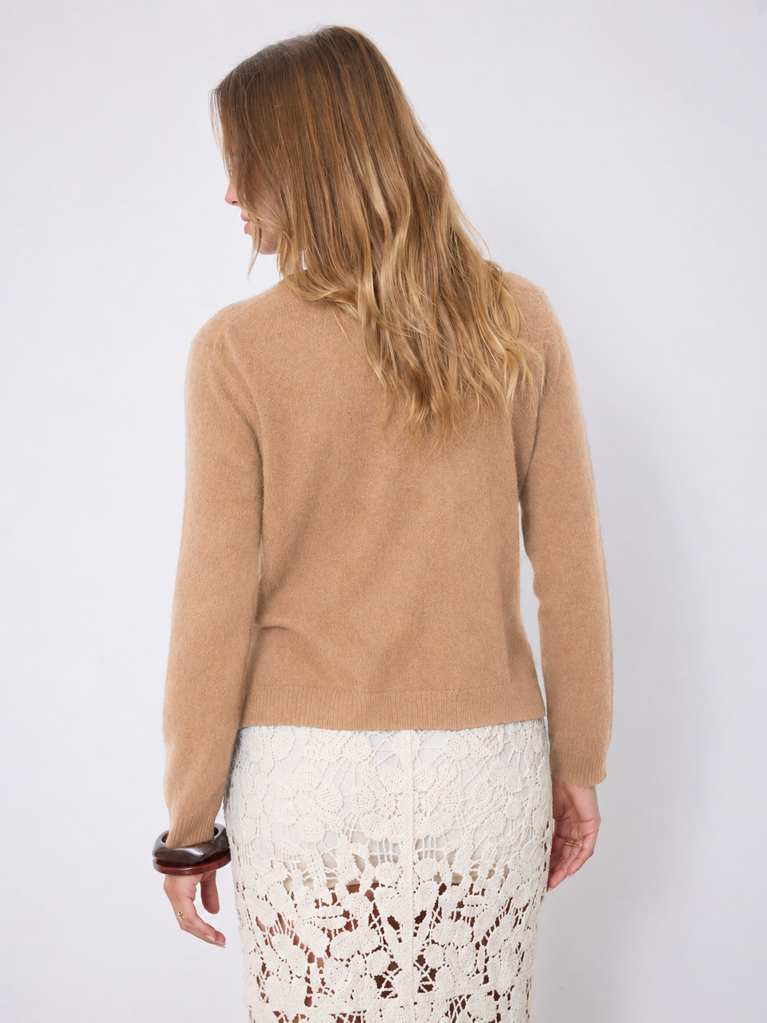 A woman shown wearing a cropped, long-sleeve knit cardigan in a warm camel tan color over a white lace skirt, set against a plain light gray studio background.
