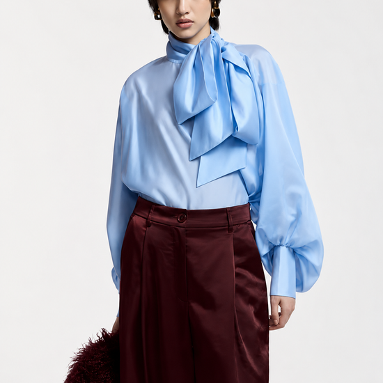 A model wearing a light blue long-sleeve blouse with a large ruffled bow at the neck and dark burgundy wide-leg trousers, holding a dark brown furry accessory against a plain light gray background.