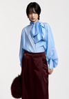 A model wearing a light blue long-sleeve blouse with a large ruffled bow at the neck and dark burgundy wide-leg trousers, holding a dark brown furry accessory against a plain light gray background.