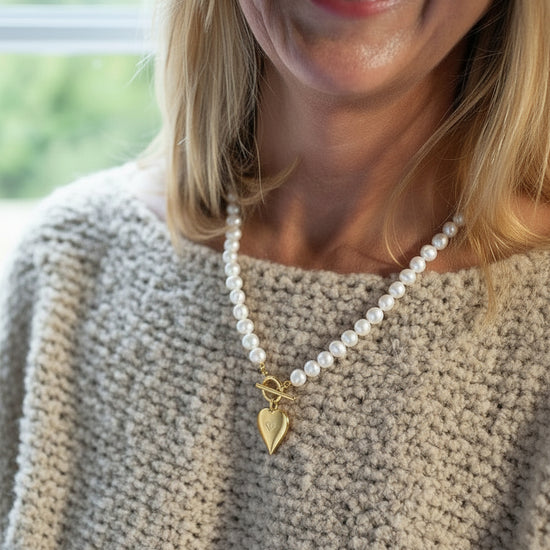 Woman wearing a pearl necklace with a heart charm, smiling outdoors.