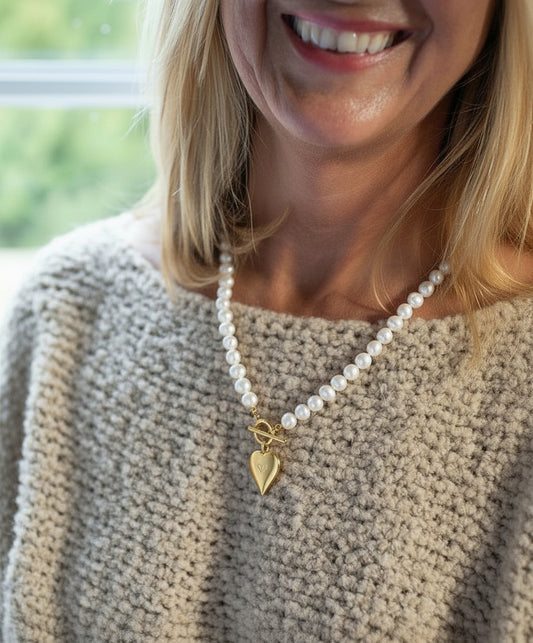 Woman wearing a pearl necklace with a heart charm, smiling outdoors.