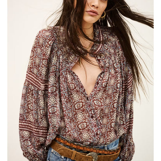 A woman wearing a bohemian-style long-sleeve blouse with a brown, red, and beige geometric pattern, paired with blue jeans and a brown belt, against a plain off-white background.