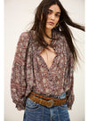 A woman wearing a bohemian-style long-sleeve blouse with a brown, red, and beige geometric pattern, paired with blue jeans and a brown belt, against a plain off-white background.