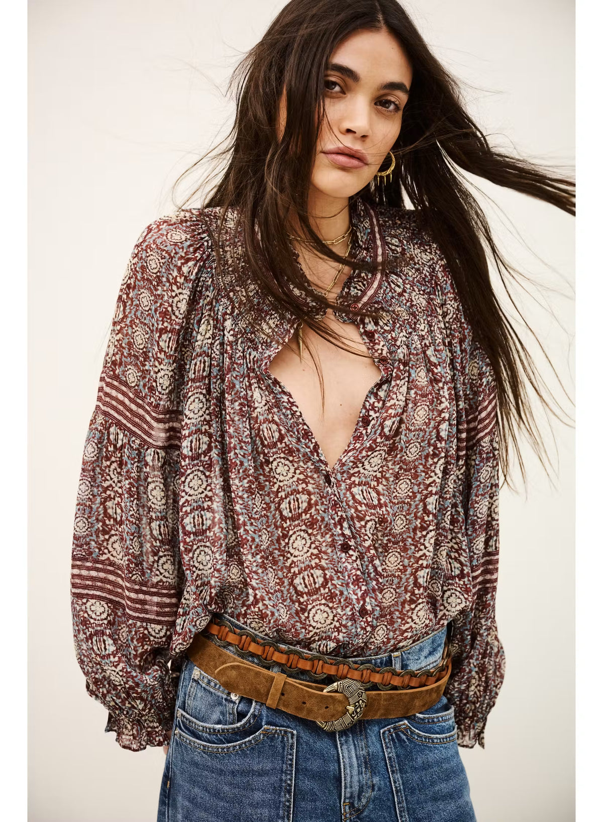 A woman wearing a bohemian-style long-sleeve blouse with a brown, red, and beige geometric pattern, paired with blue jeans and a brown belt, against a plain off-white background.