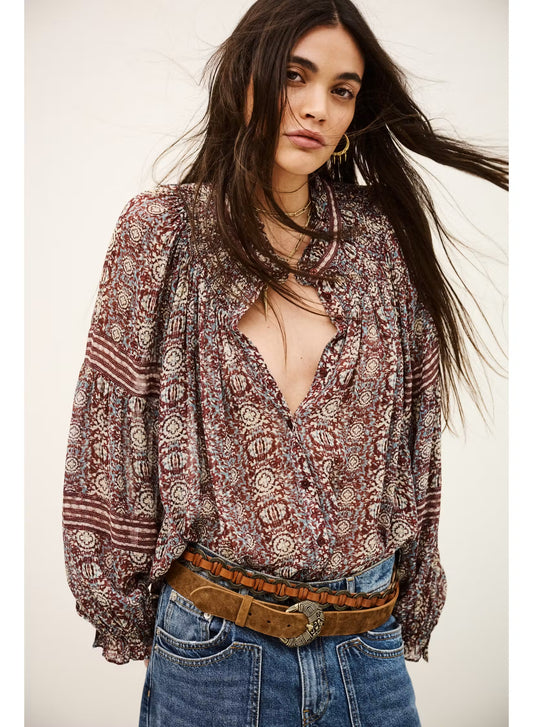 A woman wearing a bohemian-style long-sleeve blouse with a brown, red, and beige geometric pattern, paired with blue jeans and a brown belt, against a plain off-white background.