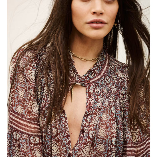 A woman wearing a bohemian-style long-sleeve blouse with a deep V-neckline and tie-neck detail, featuring a rust red, beige, and blue paisley pattern.