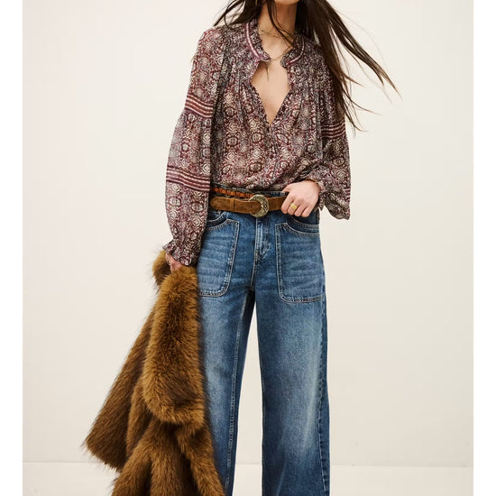 A woman stands against a plain light beige studio background wearing a long-sleeve bohemian-style patterned blouse, wide-leg blue jeans, a brown belt, and holding a brown faux fur coat.