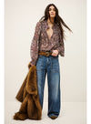 A woman stands against a plain light beige studio background wearing a long-sleeve bohemian-style patterned blouse, wide-leg blue jeans, a brown belt, and holding a brown faux fur coat.