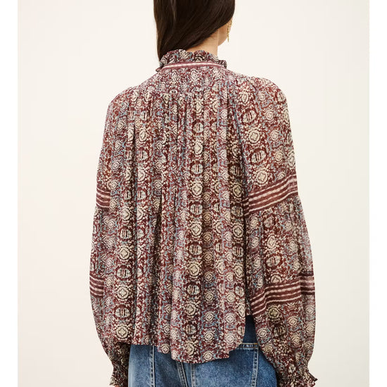 A woman viewed from the back wearing a long-sleeve bohemian patterned blouse with blue jeans, set against a plain off-white background.