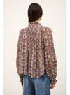 A woman viewed from the back wearing a long-sleeve bohemian patterned blouse with blue jeans, set against a plain off-white background.