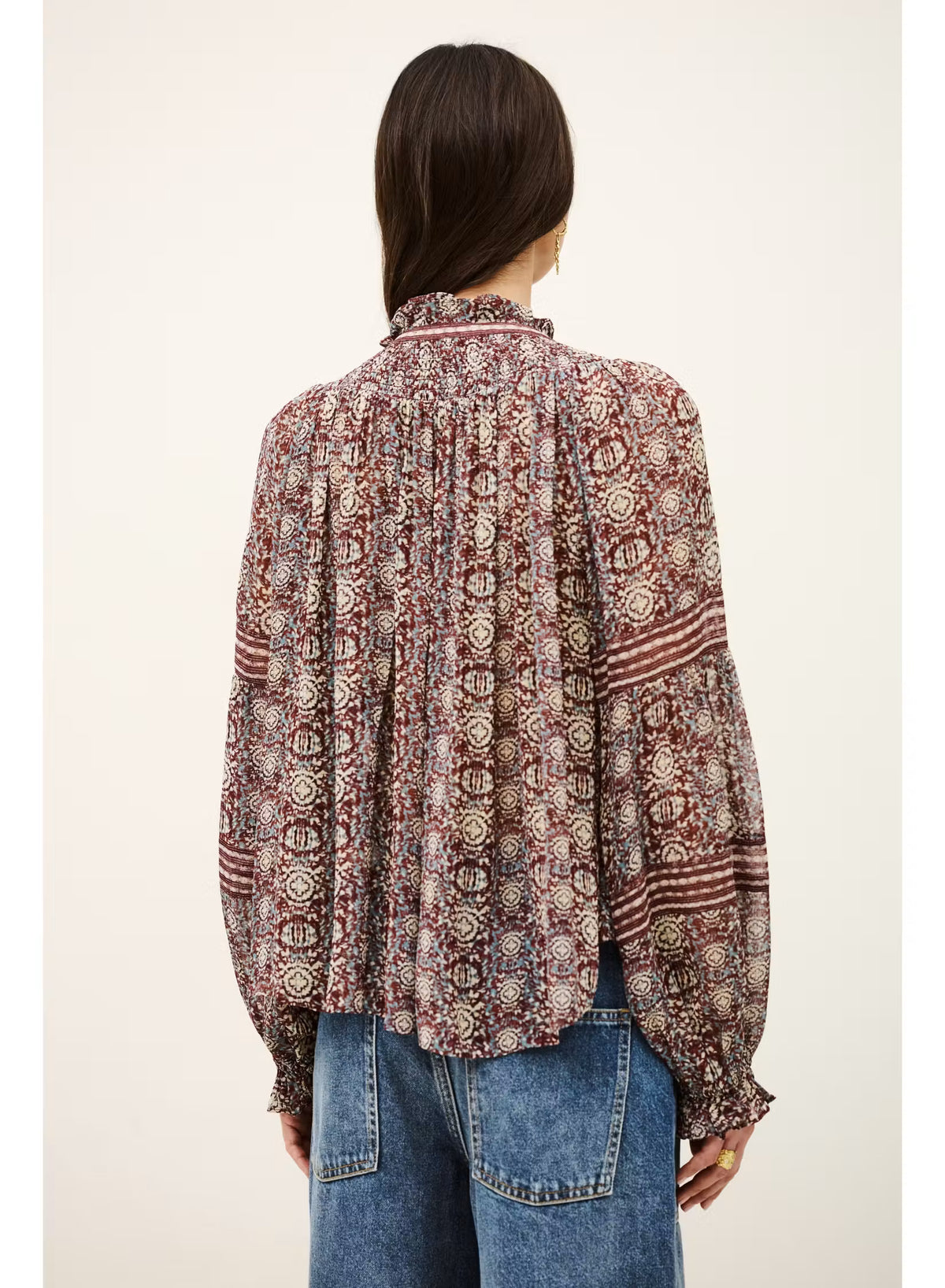 A woman viewed from the back wearing a long-sleeve bohemian patterned blouse with blue jeans, set against a plain off-white background.