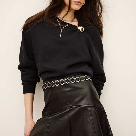 A woman modeling a black long-sleeve knit top and a black pleated faux-leather mini skirt with a silver chain-link belt, set against a plain off-white background.