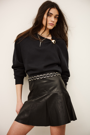A woman modeling a black long-sleeve knit top and a black pleated faux-leather mini skirt with a silver chain-link belt, set against a plain off-white background.