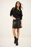 A model wearing a black crewneck sweatshirt, a black faux-leather mini skirt with a silver chain belt, and black leather ankle boots, standing against a plain off-white studio background.