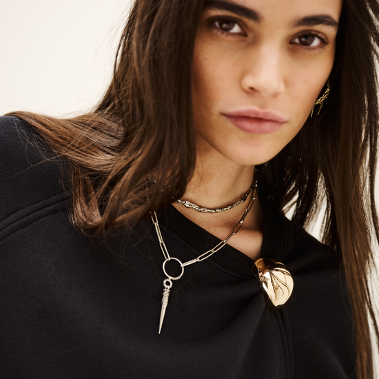 A close-up portrait of a woman with long dark brown hair wearing layered silver and gold necklaces over a black top, set against a plain light background.