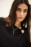 A close-up portrait of a woman with long dark brown hair wearing layered silver and gold necklaces over a black top, set against a plain light background.