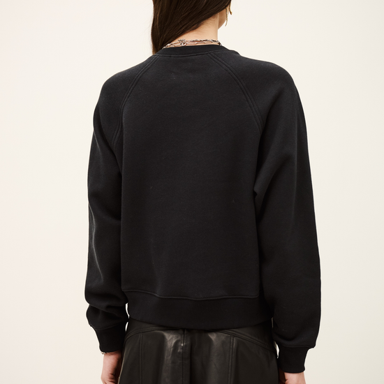 A woman shown from the back wearing a black crewneck sweatshirt and a black pleated mini skirt against a plain off-white studio background.