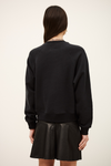 A woman shown from the back wearing a black crewneck sweatshirt and a black pleated mini skirt against a plain off-white studio background.