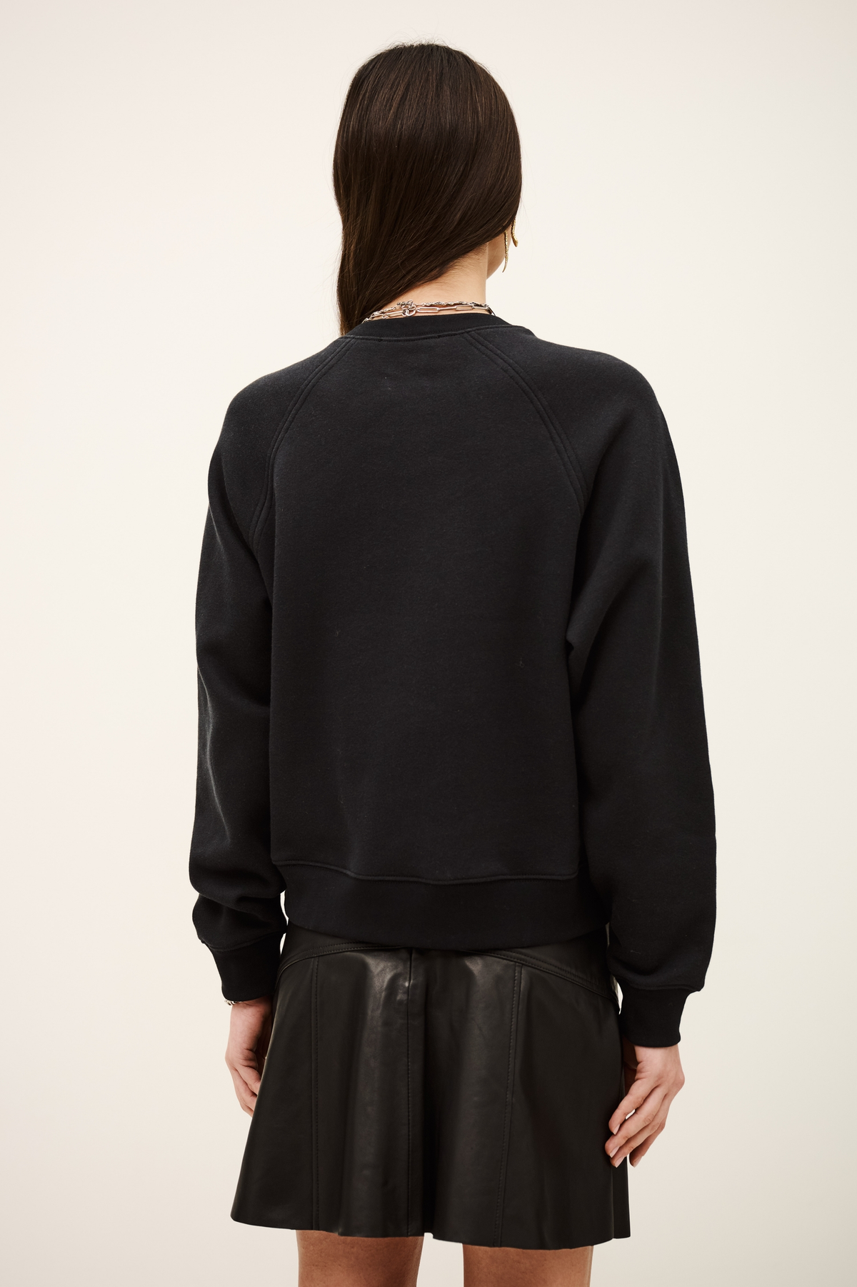 A woman shown from the back wearing a black crewneck sweatshirt and a black pleated mini skirt against a plain off-white studio background.