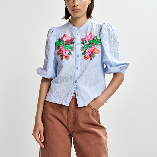 Woman wearing a light blue embroidered shirt with floral designs by Essentiel Antwerp and brown pants on a white background