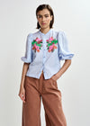 Woman wearing a light blue embroidered shirt with floral designs by Essentiel Antwerp and brown pants on a white background