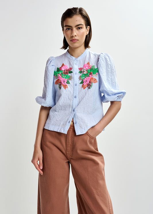 Woman wearing a light blue embroidered shirt with floral designs by Essentiel Antwerp and brown pants on a white background