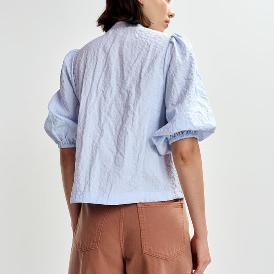 Woman wearing a light blue embroidered shirt with floral designs by Essentiel Antwerp and brown pants on a white background rear view