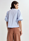 Woman wearing a light blue embroidered shirt with floral designs by Essentiel Antwerp and brown pants on a white background rear view