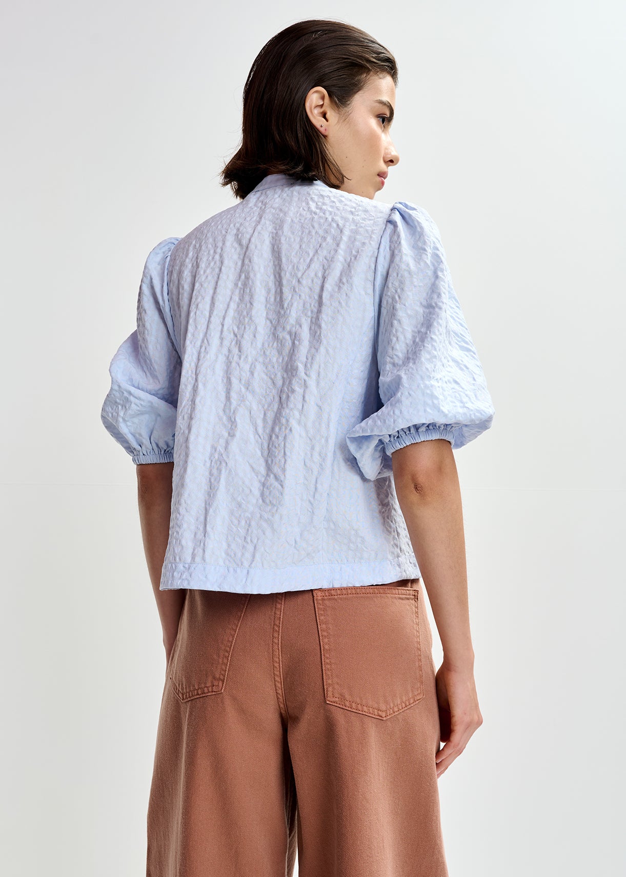 Woman wearing a light blue embroidered shirt with floral designs by Essentiel Antwerp and brown pants on a white background rear view