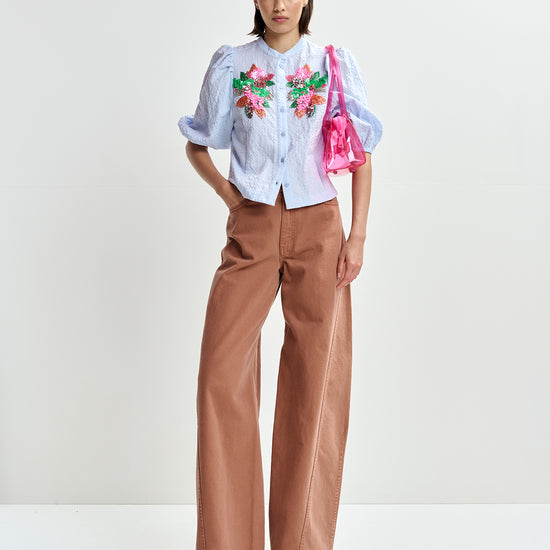 Woman wearing a light blue embroidered shirt with floral designs by Essentiel Antwerp and brown pants on a white background full body shot