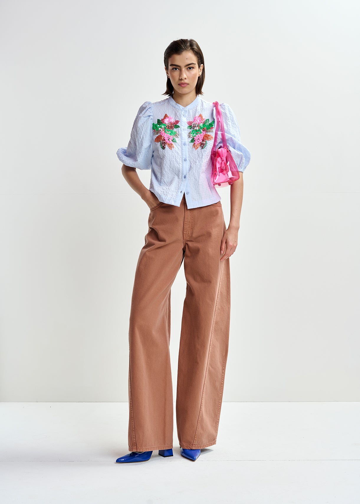 Woman wearing a light blue embroidered shirt with floral designs by Essentiel Antwerp and brown pants on a white background full body shot